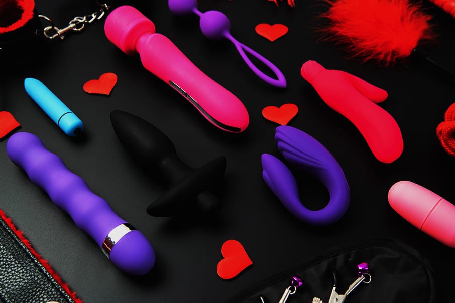 Een assortiment van kleurrijke sextoys, waaronder vibrators, buttplugs en bondageaccessoires, uitgestald op een zwarte achtergrond met rode hartjes